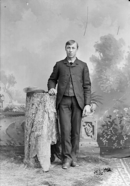 Full-length studio portrait in front of a painted backdrop of a young European American man posed standing on fake grass with his right hand resting on a tree stump. A hat is resting on top of the tree stump. He is wearing a dark-colored suit coat, vest, trousers, necktie, and watch chain.