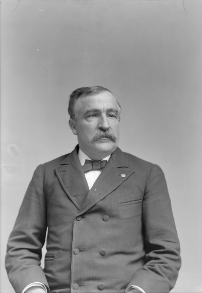 Waist-up studio portrait of a European American with a moustache posed sitting. He is wearing a dark-colored double-breasted suit coat, bow tie, and lapel pin.