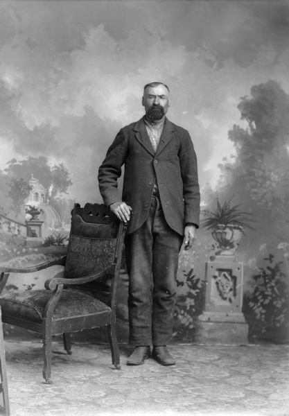 Full-length studio portrait in front of a painted backdrop of a European American man with a beard posed standing with his right hand on the back of a chair. He is wearing a dark-colored suit coat, trousers, and checked shirt.