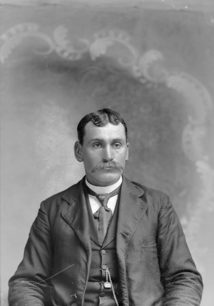Waist-up studio portrait of a European American man with a moustache posed sitting. He is wearing a dark-colored suit coat, vest, necktie, watch chain, and a clover-shaped tie pin.