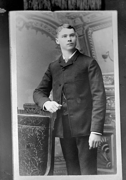 Copy photograph of a studio portrait of an unidentified European American man posed standing with his right arm resting on the back of a chair. He is wearing a dark-colored suit coat, vest, striped necktie, and watch chain.