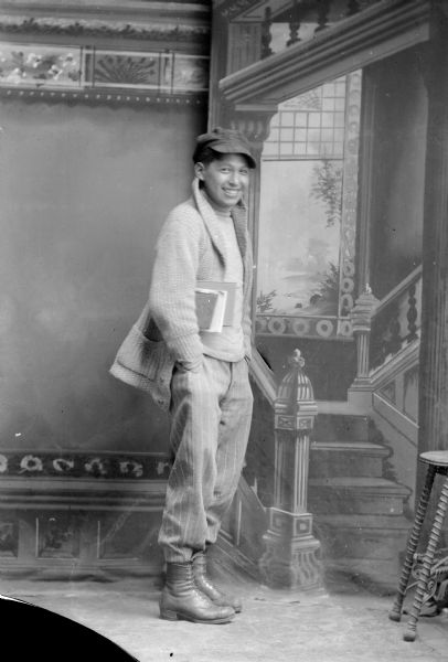 Full-length studio portrait of a smiling boy, possibly Ho Chunk, posed standing with books under his right arm with his hands in his trouser pockets. He is wearing a long sweater, striped short-cut trousers, turtleneck sweater, and boots.