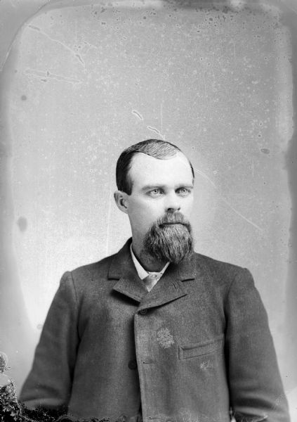 Studio portrait of a European American man with a beard posed sitting. He is wearing a dark-colored suit coat and necktie.