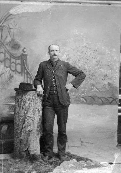 Studio portrait of a European American man with a moustache posed standing with his right arm resting on a tree stump that also holds a hat, and his left hand on his hip. He wears dark-colored trousers, suit coat, necktie, vest, and a watch chain.