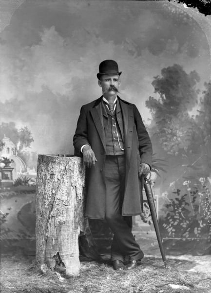Full-length studio portrait in front of a painted backdrop of a European American man with a moustache posed standing with his right arm resting on a tree stump. He is holding a rolled umbrella his left hand. He is wearing a dark-colored three-quarter length overcoat over a suit coat, vest, and necktie, and is also wearing a hat, gloves and a watch chain.