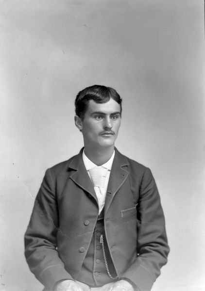 Waist-up studio portrait of an unidentified European American man with a moustache posed sitting with his hands on his lap and wearing a dark-colored suit coat, vest, watch chain, and light-colored necktie.