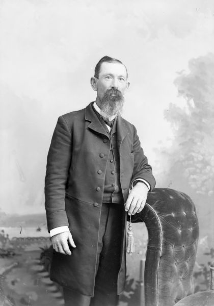 Three-quarter length studio portrait of an unidentified European American man with a beard posed standing with his left arm on the back of an overstuffed chair. He is wearing a long dark-colored suit coat, vest, and bow tie.