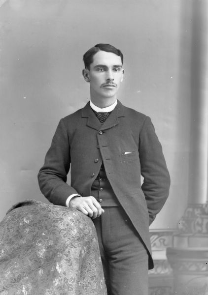 Three-quarter length tudio portrait of an unidentified young European American man posed standing. He has his left hand resting on a chair draped with a fabric, and is wearing a long dark-colored suit coat, vest, and neck tie.