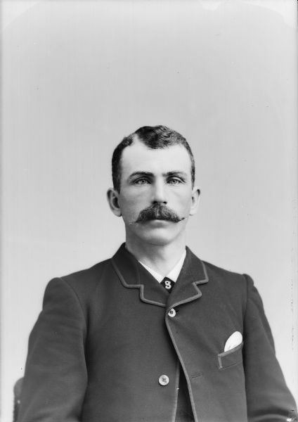 Waist-up studio portrait of an unidentified European American man with a moustache posed sitting. He is wearing a dark-colored suit coat, vest, necktie, and tie pin.