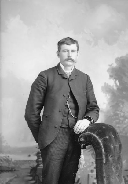 Three-quarter length studio portrait of an unidentified European American man with a moustache posed standing. He has his left arm resting on the back of an overstuffed chair, and is wearing a dark-colored suit coat, vest, and necktie.