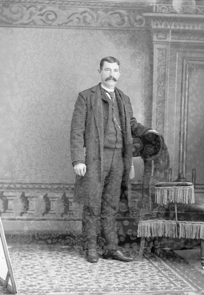 Full-length studio portrait in front of a painted backdrop of an unidentified European American man with a moustache posed standing. He has his left hand resting on the back of a chair with tassels, and is holding a hat. He is wearing a dark-colored overcoat, suit coat, vest, and necktie.