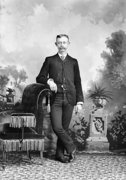 Full-length studio portrait in front of a painted backdrop of an unidentified European American man with a moustache posed standing. He has his right arm resting on the curved back of an overstuffed chair and his right leg crossed over his left leg. He wears a dark-colored suit coat, vest, striped trousers, necktie, and watch chain.