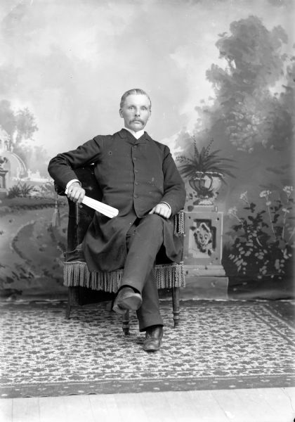 Full-length studio portrait in front of a painted backdrop of an unidentified European American man with a moustache holding a rolled-up  newspaper in his right hand. He is posed sitting in a tasseled arm chair with his left leg crossed over his right leg. He is wearing a dark-colored buttoned-up overcoat.