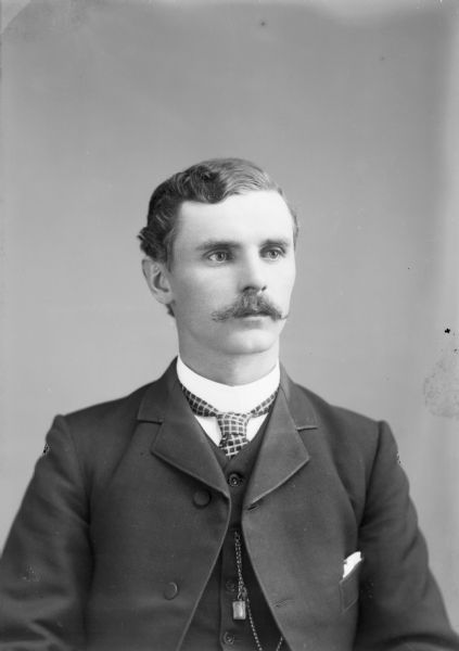 Quarter-length studio portrait of an unidentified European American man with a moustache posed sitting. He is wearing a dark-colored suit coat, vest, watch chain fob, and plaid neck tie.