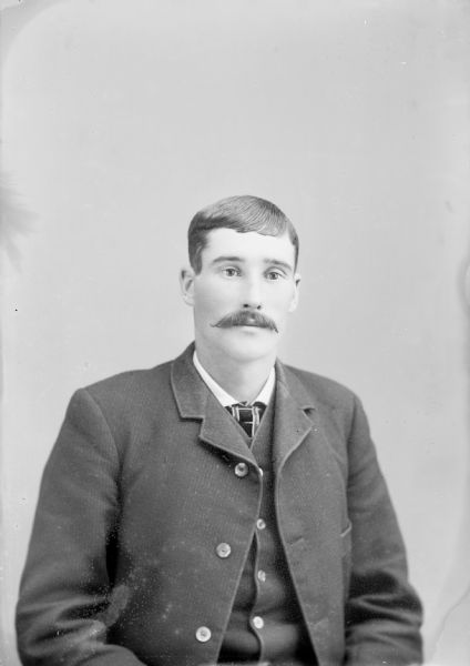 Waist-up studio portrait of an unidentified European American man with a moustache posed sitting. He is wearing a dark-colored suit coat, vest, and striped neck tie.