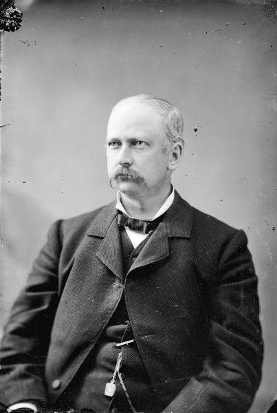 Waist-up studio portrait of an unidentified European American man with a moustache posed sitting. He is wearing a dark-colored suit coat, vest, watch chain, and bow tie.