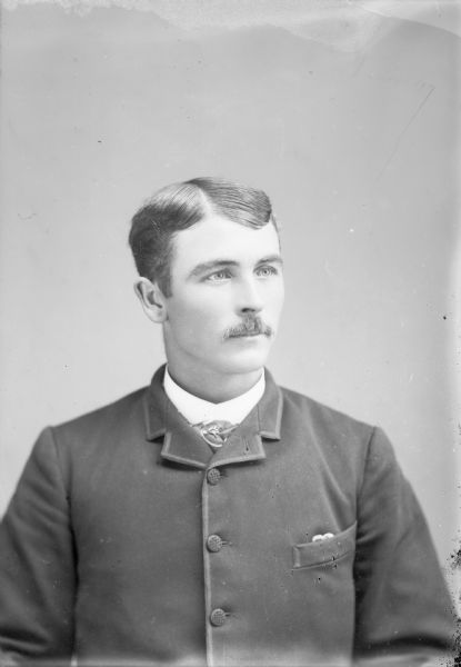 Waist-up studio portrait of an unidentified young European American man posed sitting. He is wearing a dark-colored suit coat, vest, watch chain, and light-colored necktie.