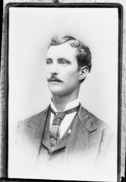 Copy photograph of a studio portrait of an unidentified European American man with a moustache. He is wearing a dark-colored suit coat, vest, neck tie, tie pin, and lapel pin.