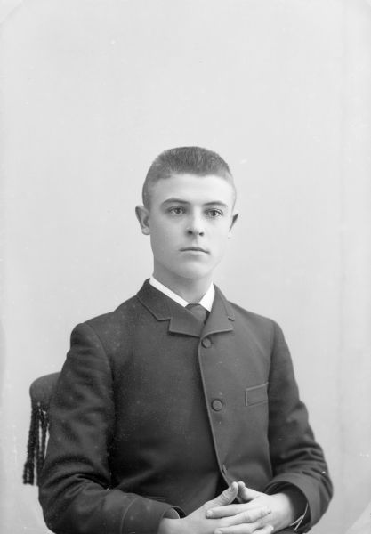 Studio portrait of an unidentified young European American man posed sitting with his hands clasped. He is wearing a dark-colored button-up suit coat and a neck tie.