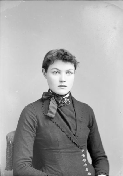 Waist-up studio portrait of an unidentified European American woman posed sitting. She is wearing a dark-colored dress with a v-shaped neckline, buttons in the front, a ribbon around from her neck, and a collar pin.
