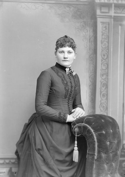 Studio portrait of an unidentified European American woman posed standing with her hands on the back of an overstuffed chair, and wearing a dark-colored dress with a button-down front, embroidered decorations, and a collar brooch.