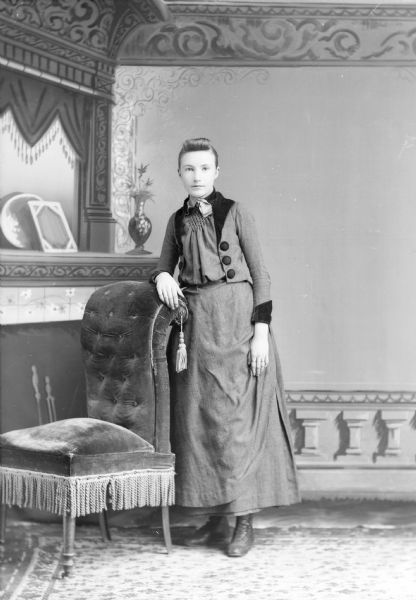 Full-length studio portrait in front of a painted backdrop of an unidentified European American woman posed standing with her right arm resting on the curved back of an overstuffed chair with tassels. She is wearing a dark-colored dress with a darker-colored trim, a ribbon around her neck, and a collar pin.