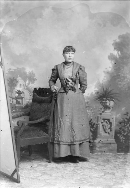 Studio portrait in front of a painted backdrop of an unidentified European American woman posed standing with her right hand on the back of an overstuffed chair. She is wearing a dark-colored dress with a bow at the waist. A light reflecting board is visible on the left.