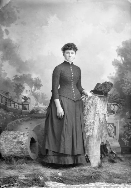 Full-length studio portrait in front of a painted backdrop of an unidentified European American woman posed, standing on fake grass on top of a carpet, and resting her left arm on a stump, next to a feathered hat. She is wearing a dark-colored skirt, button-down jacket with coin buttons, earrings, and a collar pin.