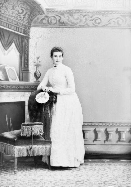 Full-length studio portrait in front of a painted backdrop of an unidentified European American woman posed standing. She is resting both arms on the curved back of a tasseled, overstuffed chair, and is holding a hand fan in her right hand. She is wearing a light-colored dress with a button-down bodice and a collar pin.