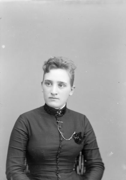 Waist-up studio portrait of an unidentified European American woman posed sitting. She is wearing a dark-colored dress with a button-down bodice, watch chain, and a collar pin. The watch is inside a pocket over her left breast.