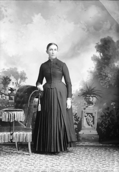 Full-length studio portrait in front of a painted backdrop of an unidentified European American woman posed standing with her right hand on the curved back of an overstuffed chair with tassels. She is wearing a dark-colored dress with a button-down bodice, a pleated skirt and a collar pin.
