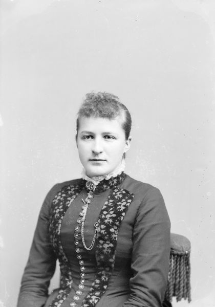 Waist-up studio portrait of an unidentified European American woman sitting in a chair with tasseled fringe on the back. She is wearing a dark-colored dress with coin buttons, a lace ruffle at the collar, and floral print trim. She also wears earrings, a collar pin, and a watch chain.
