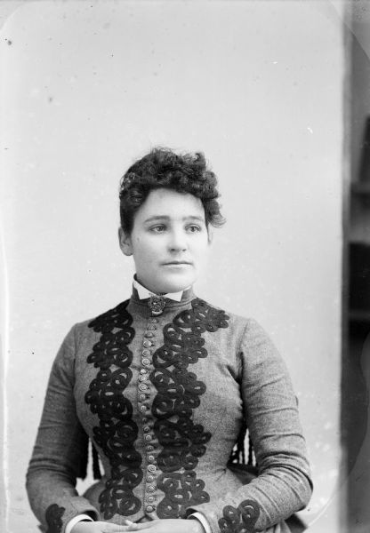 Waist-up studio portrait of an unidentified European American woman posed sitting in a chair with tasseled trim on the back. She is wearing a light-colored jacket with metal buttons and dark-colored appliquéd spiral trim, and a collar pin.