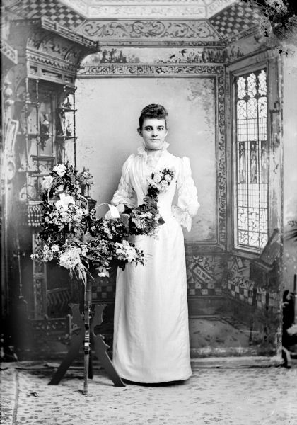 Full-length studio portrait in front of a painted backdrop of an unidentified European American woman posed standing next to a floral display on a stand. She is holding a floral bouquet in her right hand, and is wearing a light-colored dress with a ruffled bodice, lace at neck and wrists and puffed sleeves. She also wears a choker necklace and a corsage.