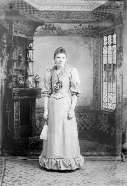 Full-length studio portrait in front of a painted backdrop of an unidentified, European American woman posed standing. She has her arms at her sides and is wearing full-length fingerless gloves and a corsage. She is holding a hand fan in her right hand and is wearing a light-colored dress with a ruffled bodice, ruffled hem and puffed sleeves.