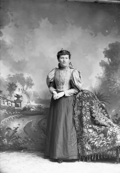Full-length studio portrait in front of a painted backdrop of an unidentified European American woman posed standing with her left arm resting on a chair draped with fabric. She is wearing a hair comb in her hair, a dark-colored skirt, a ring on her left hand, and a light-colored striped blouse with a large bow at the back of her neck.