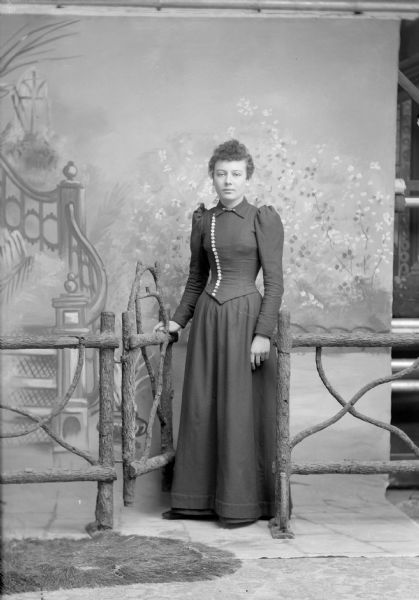 Full-length studio portrait of a European American woman posed standing in the open gateway of a prop wooden fence with her right hand on the gate. She is wearing a dark-colored dress with a button-down bodice and collar pin.