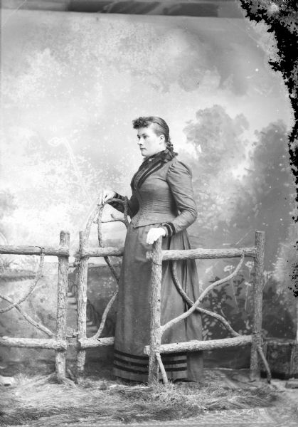 Full-length studio portrait in front of a painted backdrop of a European American woman posed standing in the open gateway of a prop wooden fence facing towards the left. She is wearing a dark-colored dress with velvet trim on the bodice, wrists and just above the hem. She is wearing a collar pin and a ribbon at her neck.