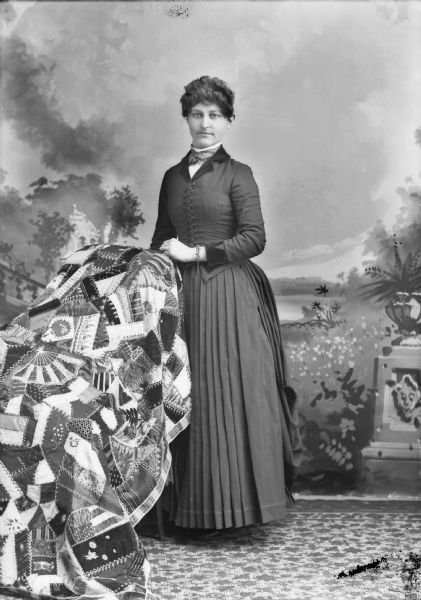 Full-length studio portrait in front of a painted backdrop of a European American woman posed standing. She is resting her left hand on the back of a chair, and is wearing a bracelet with a chain. The chair is covered with a quilt. She is wearing a dark-colored dress with a button-down bodice and pleated skirt.