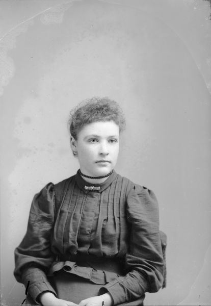 Waist-up studio portrait of a European American woman posed sitting. She is wearing a dark-colored pleated blouse, bar-shaped collar pin, and a double pearl strand choker.