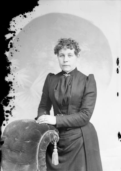 Three-quarter length studio portrait of an unidentified European American woman posed standing with her hands on the back of an overstuffed chair. She is wearing a dark-colored dress with a pleated bodice and a collar pin.