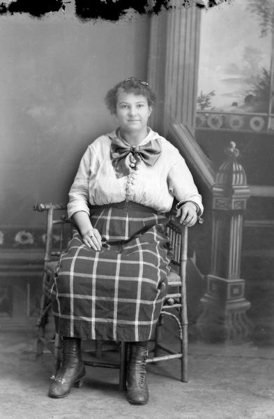 Studio portrait in front of a panted backdrop of a European American woman posed sitting in a wooden chair. She is wearing a plaid skirt and light-colored blouse with a large bow collar.