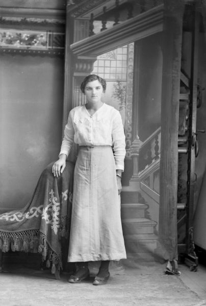 Full-length studio portrait in front of a painted backdrop of a European American woman posed standing with her right hand on the back of a chair covered with a tasseled fabric. She is wearing a comb in her hair, a bracelet, a light-colored blouse with a round pin at the collar, and a long skirt that is hemmed just above her ankles.