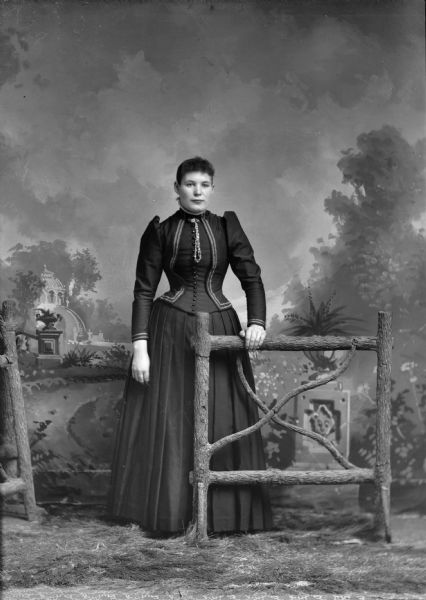 Full-length studio portrait in front of a painted backdrop of a European American woman posed standing in the gateway of a prop wooden fence. She has her left hand resting on the fence and is wearing a dark-colored dress with light-colored trim, collar pin, and buttonhole chain.