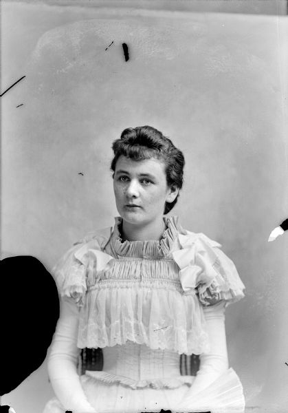 Waist-up studio portrait of a European American woman posed sitting. She is wearing a light-colored dress with a ruffled bodice and sleeves, and is holding a hand-fan.
