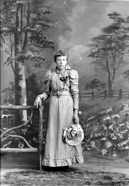 Full-length studio portrait in front of a painted backdrop of a European American woman posed standing next to a prop fence. She is wearing a light-colored dress and a corsage, and is holding a straw hat decorated with ribbons and flowers in her left hand.