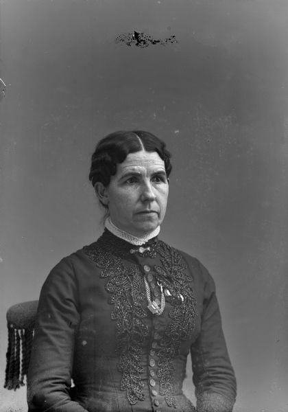Studio portrait of an unidentified European American woman posed sitting in a chair. She is wearing a dark-colored dress with embroidered decoration and metal buttons. She also has buttonhole chain with a watch tucked under the embroidery, and is wearing a collar pin.