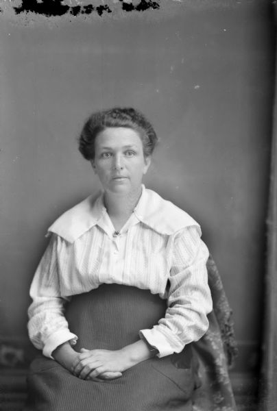 Studio portrait of an unidentified European American woman posed sitting on a cloth-covered chair with her hands clasped in her lap. She is wearing a light-colored blouse, a collar pin, and a dark-colored skirt.