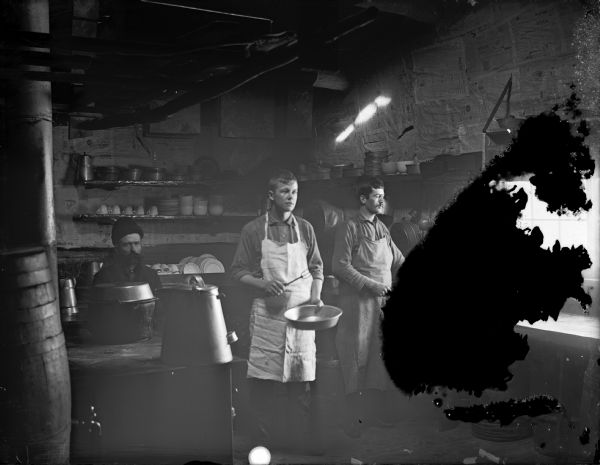 Interior view of a kitchen in a logging camp with light coming in from a window on the far right. Two men are posing standing, and another man is sitting in the back on the left.