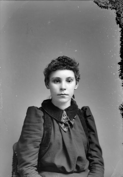 Waist-up studio portrait of an unidentified young European American woman posed sitting in a chair with a tasseled back. She is wearing a dark-colored dress with a wide velvet collar and a collar pin/chain.